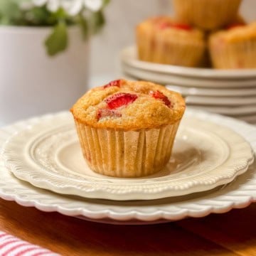 A single strawberry muffin sits atop a stack of white plates, with more delicious strawberry muffins and a potted plant softly blurred in the background.