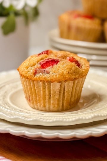 A single strawberry muffin sits atop a stack of white plates, with more delicious strawberry muffins and a potted plant softly blurred in the background.