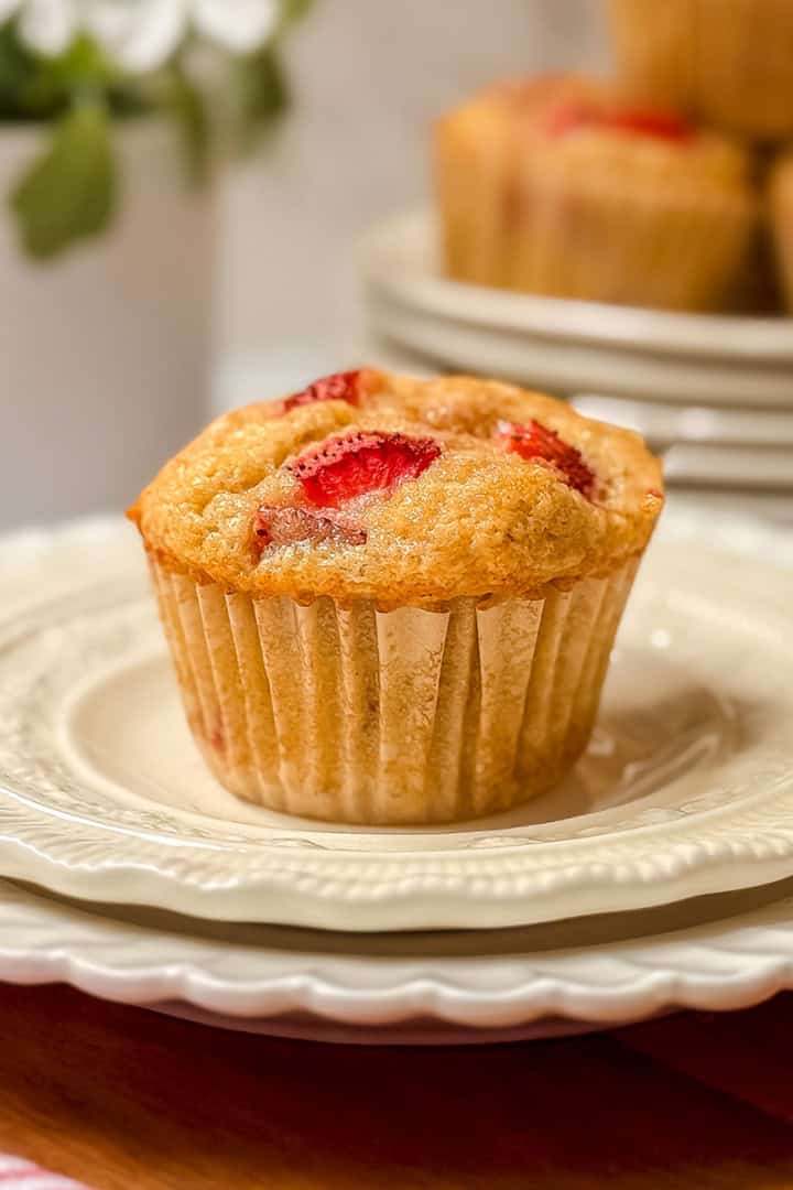 A single strawberry muffin sits atop a stack of white plates, with more delicious strawberry muffins and a potted plant softly blurred in the background.