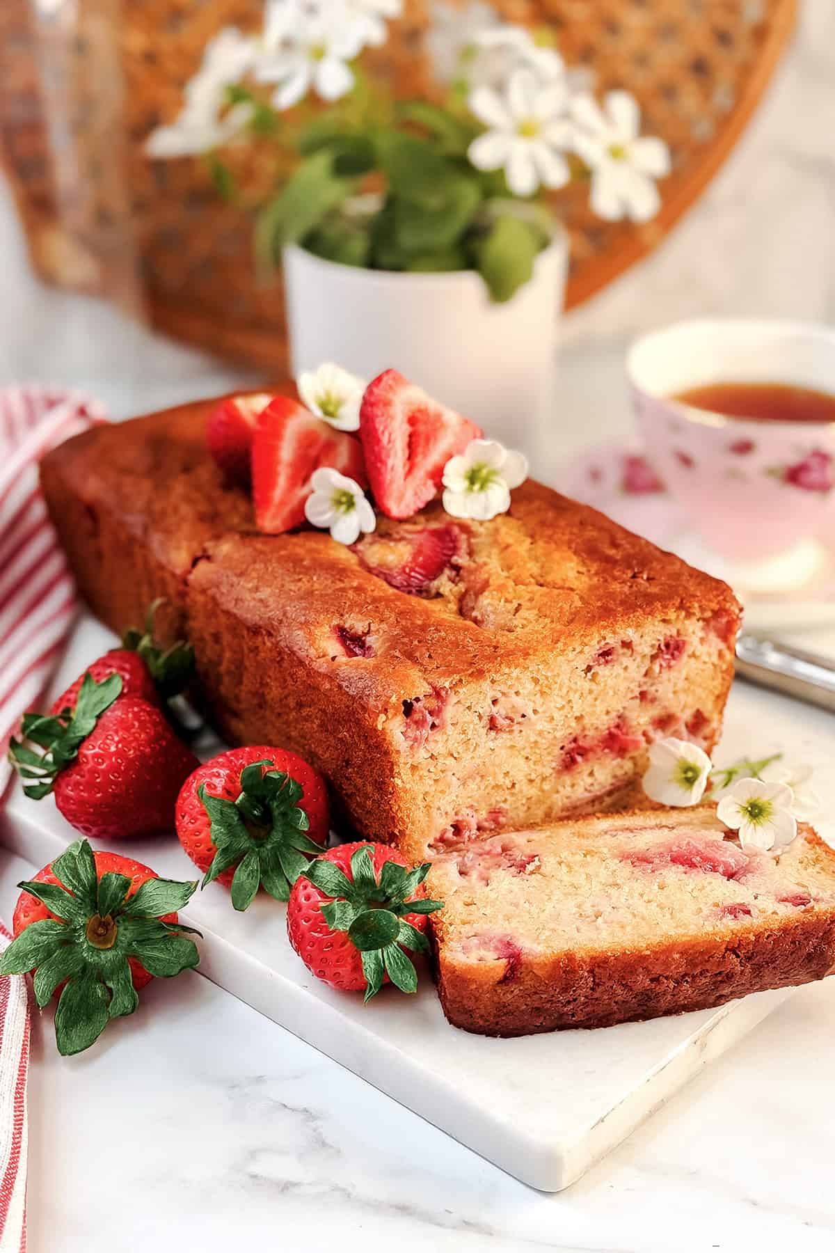 A loaf of strawberry yogurt bread topped with fresh strawberries and white flowers, with sliced strawberries and a tea cup in the background.