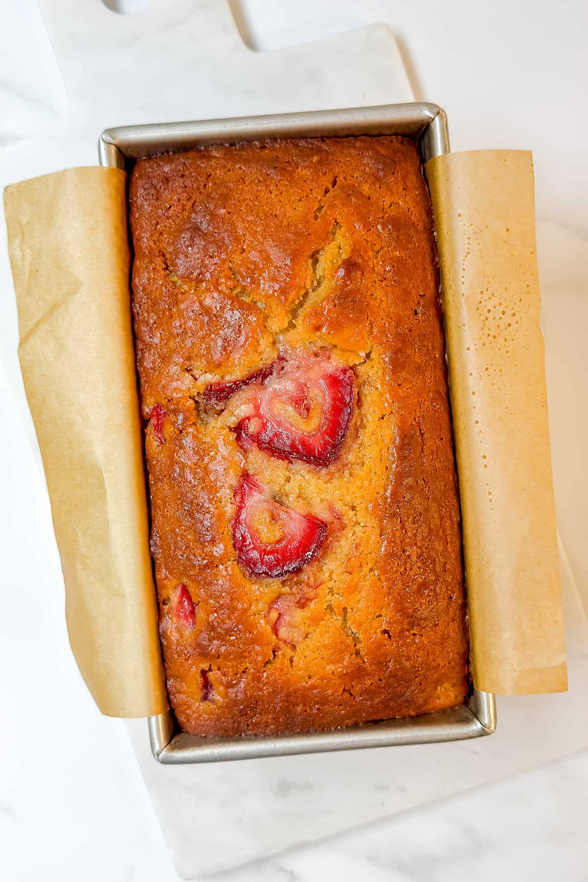 A rectangular loaf of golden-brown strawberry bread, topped with sliced strawberries, sits in a metal pan lined with parchment paper on a white surface-perfect for your next Feast Recipe Template.