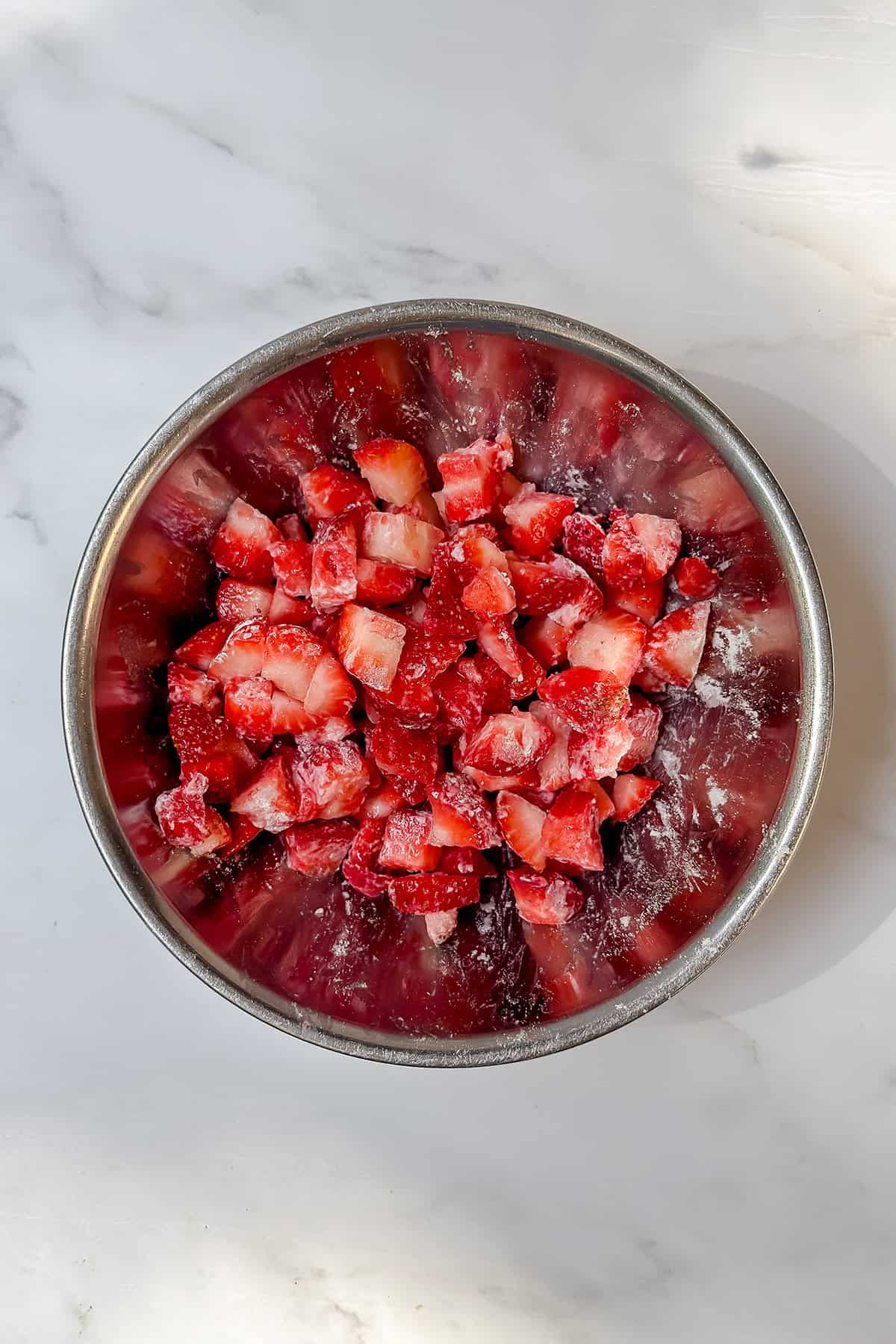 A metal bowl filled with chopped fresh strawberries coated lightly in sugar sits on a white marble surface, ready for your next Feast Recipe Template.