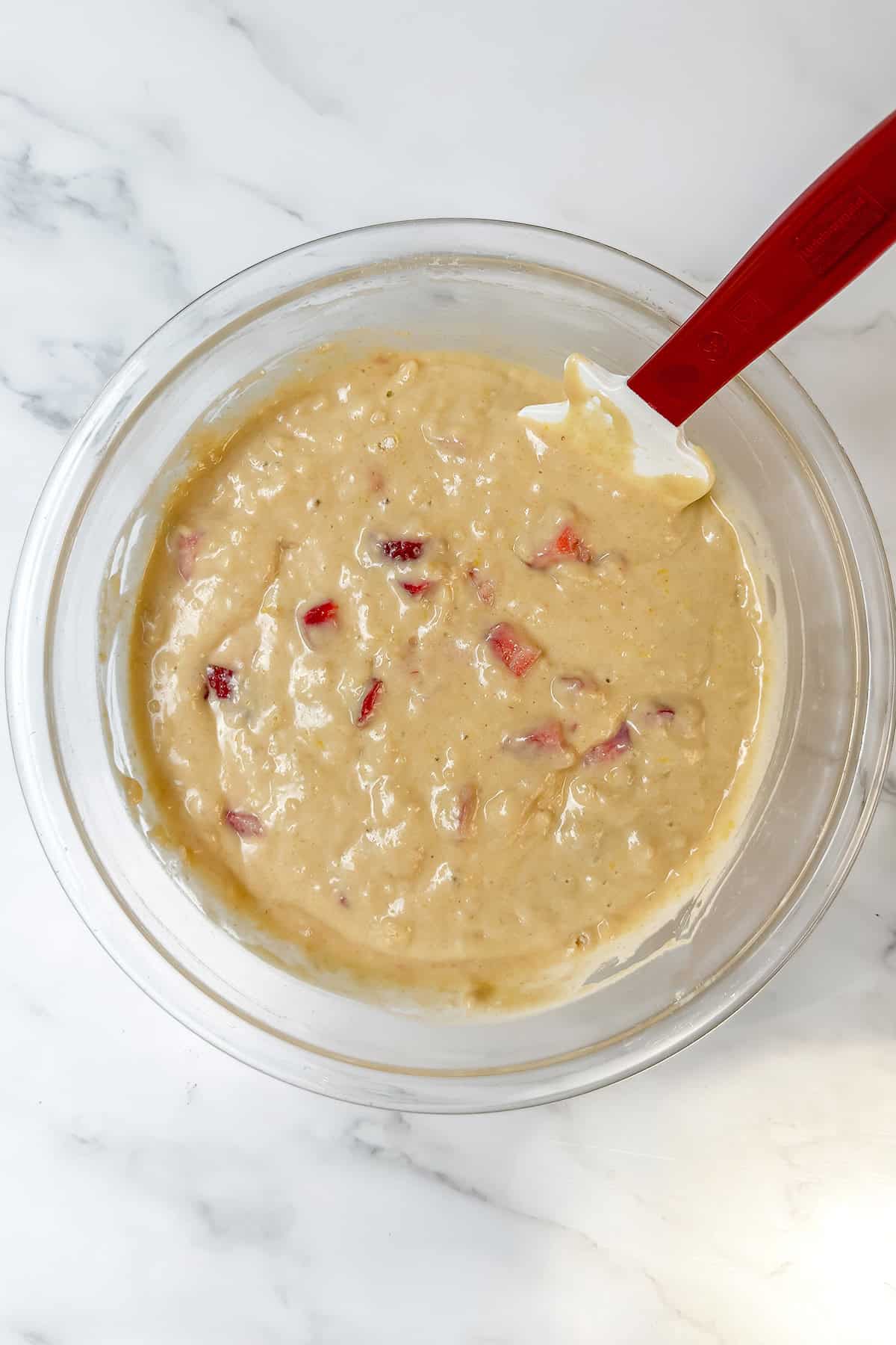 Glass bowl with pale, thick batter containing small chunks of red fruit, being mixed with a white spatula with a red handle on a marble surface-a perfect scene for your next Feast Recipe Template.