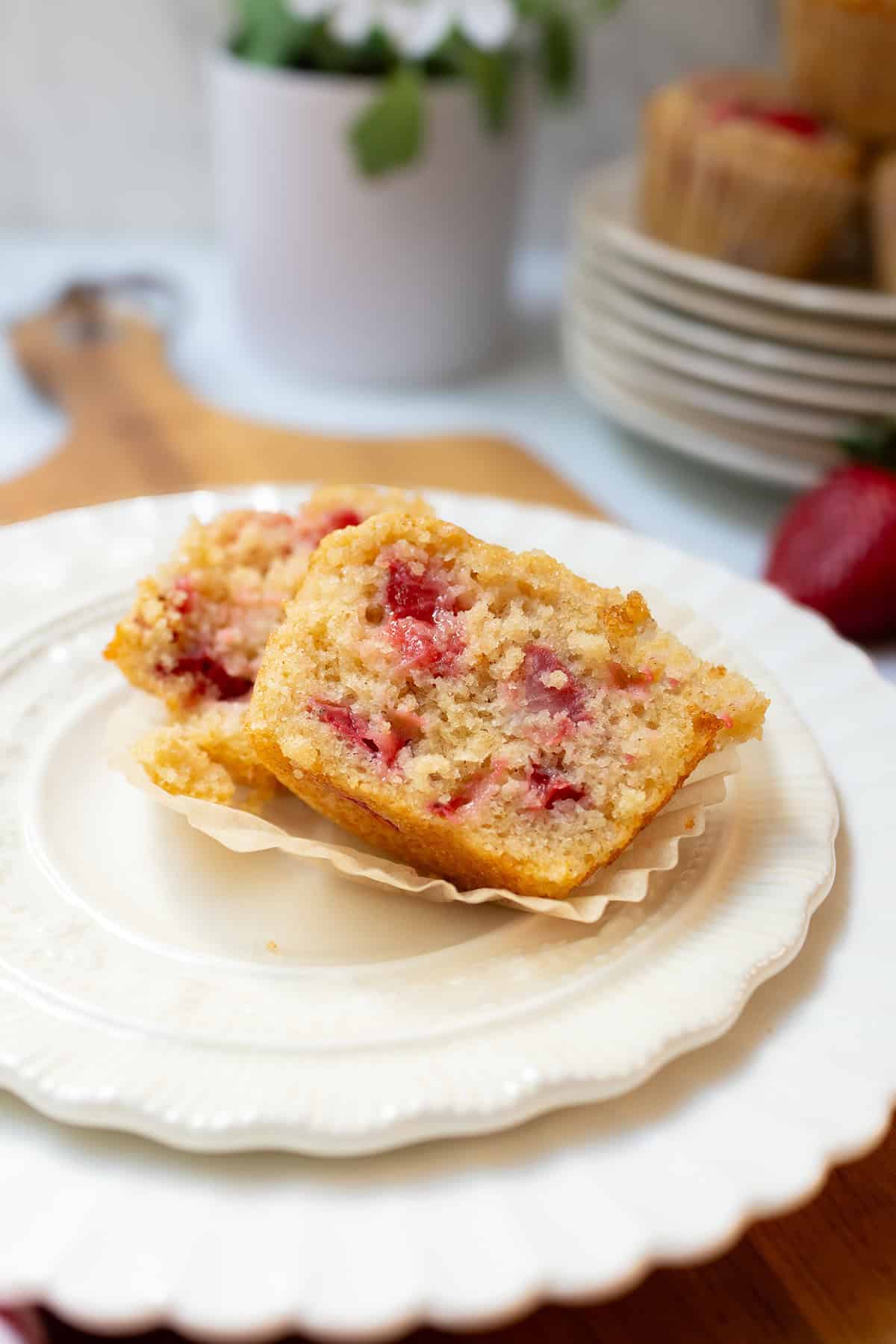 A sliced strawberry muffin sits on a white plate, revealing pieces of fresh strawberry inside. A stack of plates and a potted plant are visible in the background.