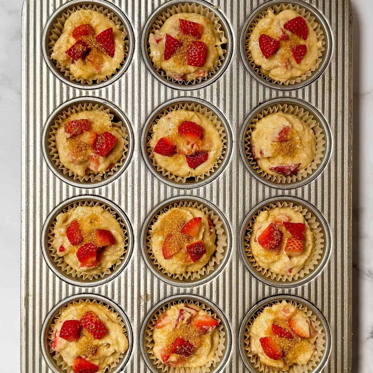 A muffin tin filled with twelve unbaked strawberry muffins, each topped with chopped strawberries and coarse sugar, set on a white marble surface.