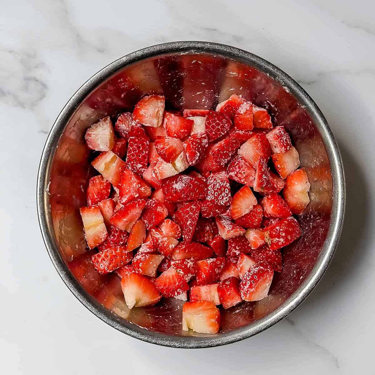 A metal bowl filled with chopped strawberries, perfect for making strawberry muffins, sits on a white marble surface.