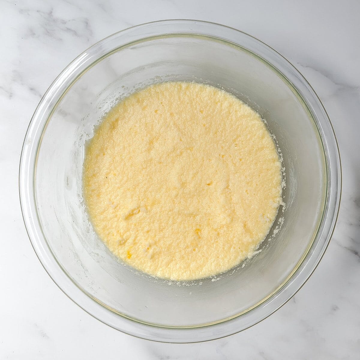 A glass mixing bowl containing a pale yellow, smooth and slightly frothy batter for Strawberry Muffins sits on a white marble surface.
