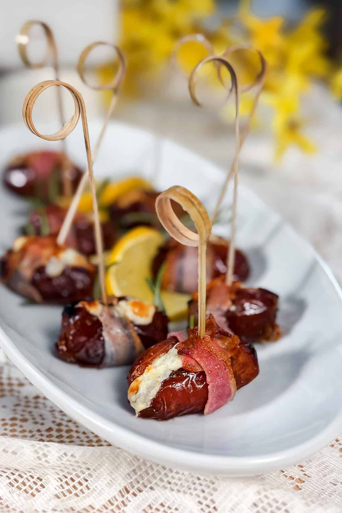A white plate with bacon-wrapped, blue cheese-stuffed dates secured with decorative toothpicks, garnished with lemon slices and herbs, makes a stunning appetizer on a lace tablecloth.