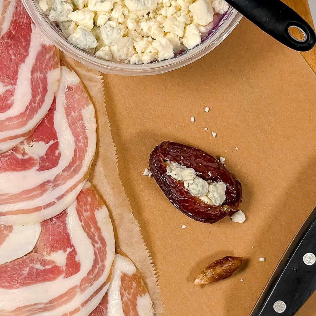 Sliced pancetta, a container of crumbled blue cheese with a measuring spoon, a stuffed date, a date pit, and a knife on a brown surface-perfect starters for your next appetizer spread.