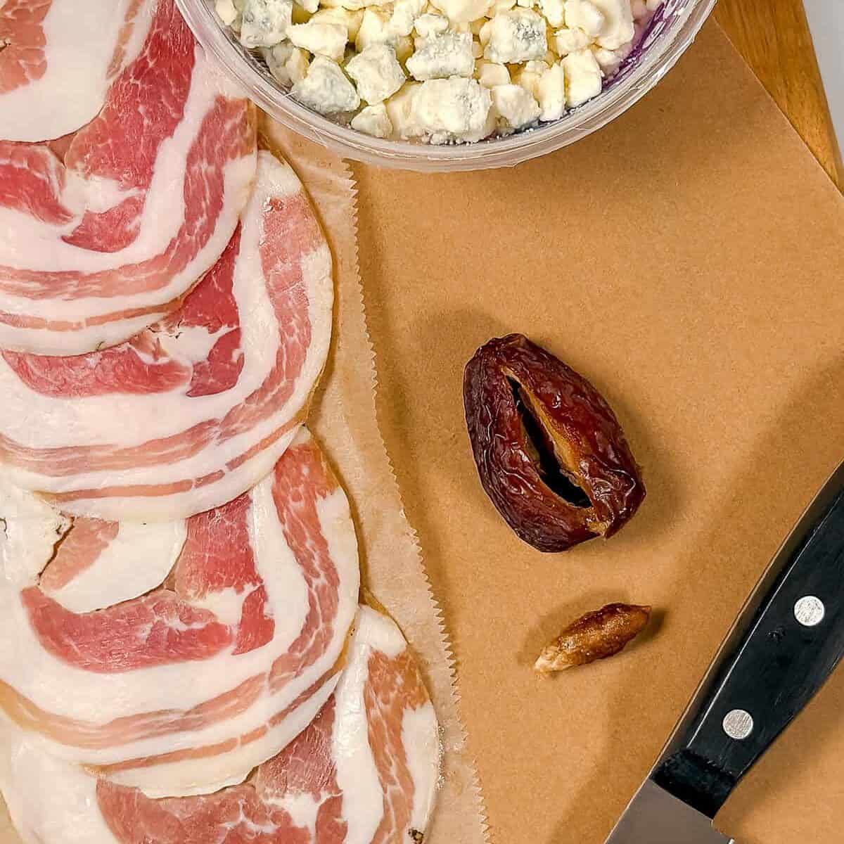 Sliced pancetta, crumbled blue cheese, a pitted date, its seed, and a knife rest on a brown cutting board-perfect for preparing blue cheese stuffed dates, an irresistible appetizer.