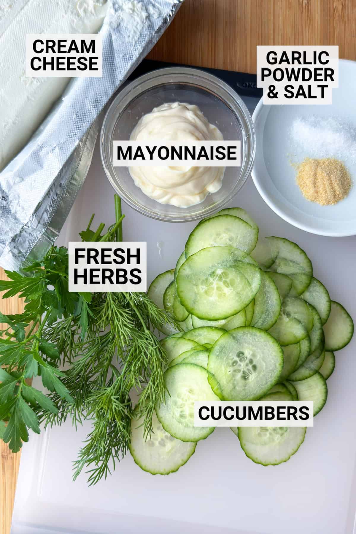 Sliced cucumbers, fresh herbs, cream cheese, mayonnaise, garlic powder, and salt-classic ingredients for Kentucky Benedictine tea sandwiches-arranged on a cutting board with each ingredient labeled.