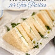 Rectangular tea sandwiches, inspired by classic Kentucky Benedictine, are filled with a creamy spread and garnished with herbs and edible flowers, arranged neatly on a white platter.
