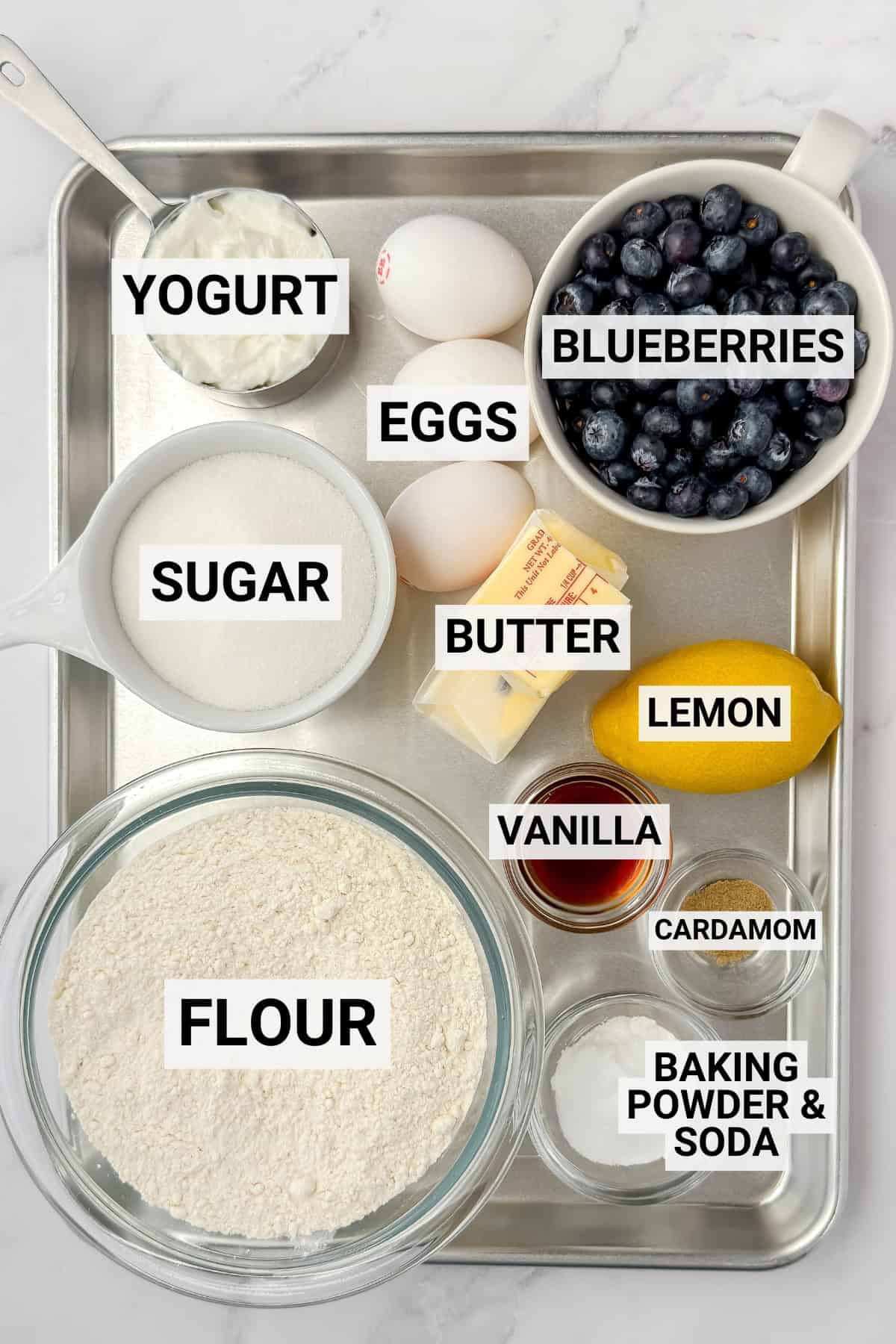 A tray with labeled ingredients for a delicious blueberry teacake: yogurt, eggs, blueberries, sugar, butter, lemon, vanilla, cardamom, flour, baking powder, and baking soda.