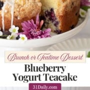Round blueberry yogurt teacake with glaze and purple flowers on top, displayed on a white cake stand with fresh blueberries and flowers around it. Text reads: "Blueberry Yogurt Teacake.