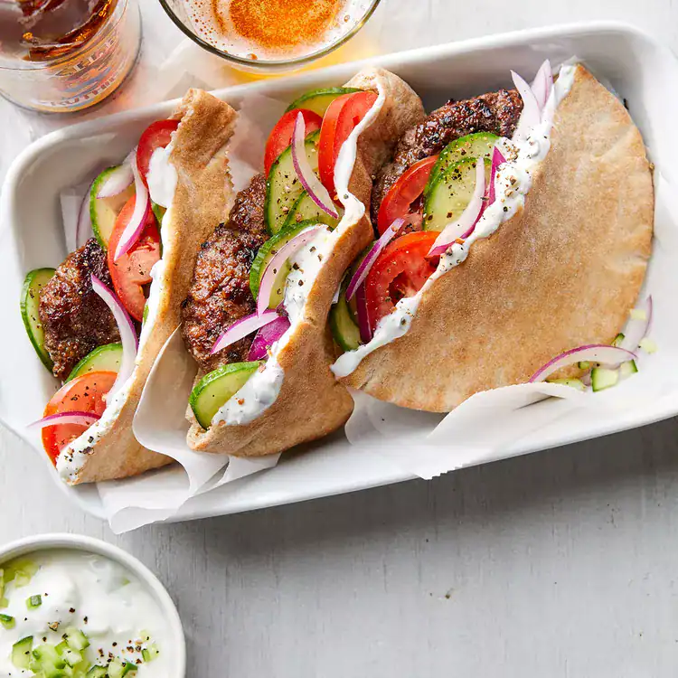 Three pita sandwiches filled with grilled meat patties, sliced cucumber, tomato, red onion, and tzatziki sauce are served on a white tray-a fresh idea for May one pot dinners-with a small bowl of extra sauce on the side.