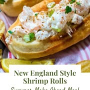 A close-up of a New England style shrimp roll with shrimp salad on a toasted bun, garnished with herbs, next to lemon slices and chips. Text: "New England Style Shrimp Rolls. Try this easy shrimp rolls recipe—your perfect summer make ahead meal.