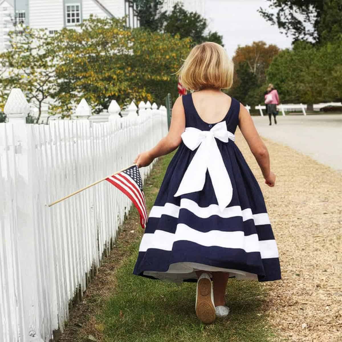 A young girl in a navy and white striped dress with a large bow walks along a path by a white fence, holding a small American flag-perfect for celebrating Mother's Day with family.