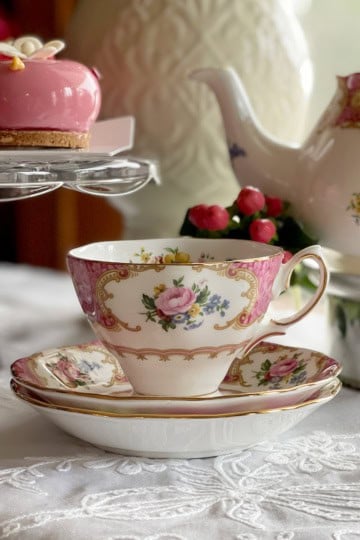 A floral teacup and saucer set on a lace tablecloth with a teapot and pink dessert nearby.
