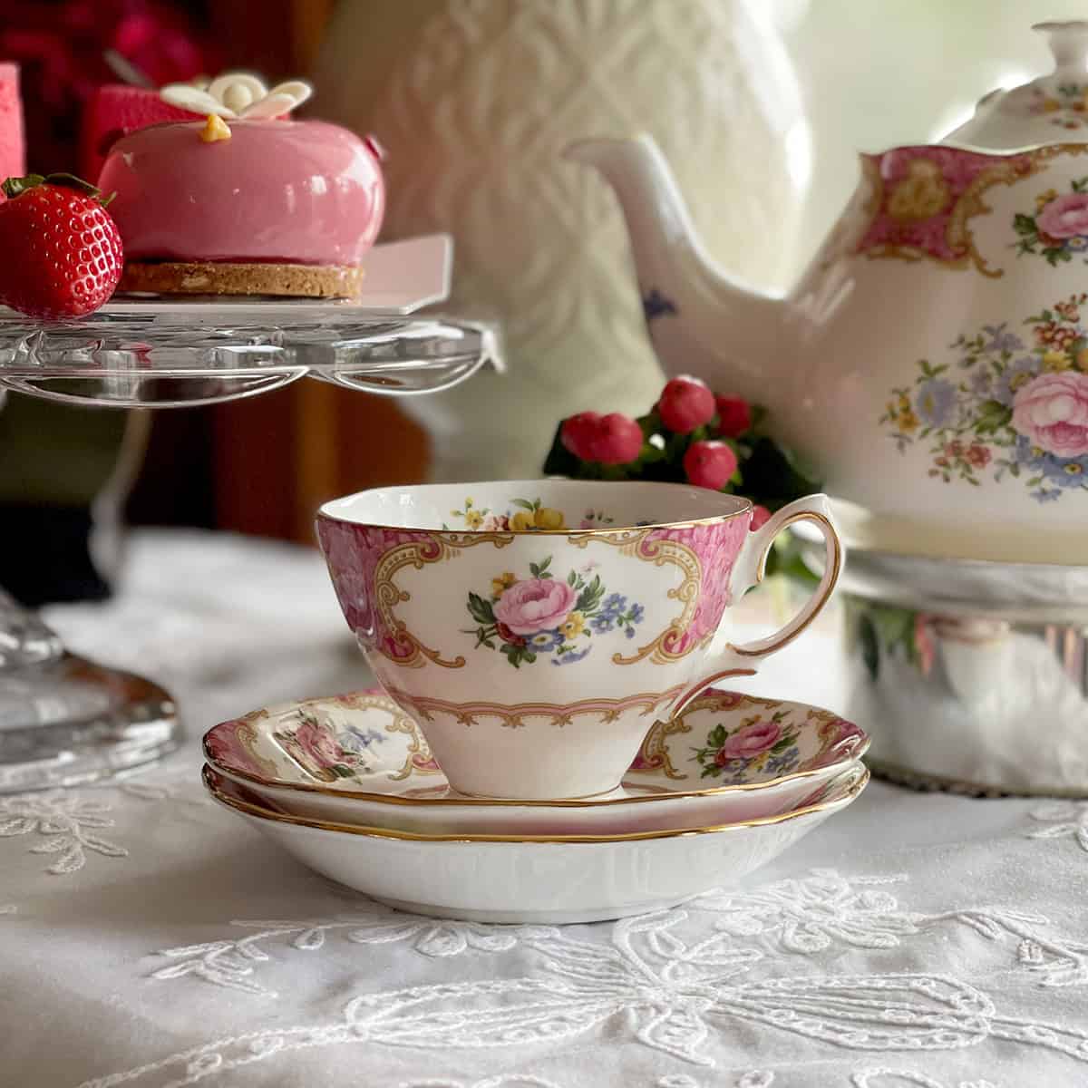 A floral teacup and saucer set on a lace tablecloth with a teapot and pink dessert nearby.