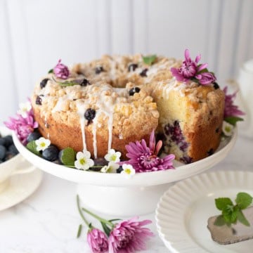 A blueberry yogurt teacake with icing is displayed on a white cake stand, garnished with fresh flowers and surrounded by plates and a bowl of blueberries.
