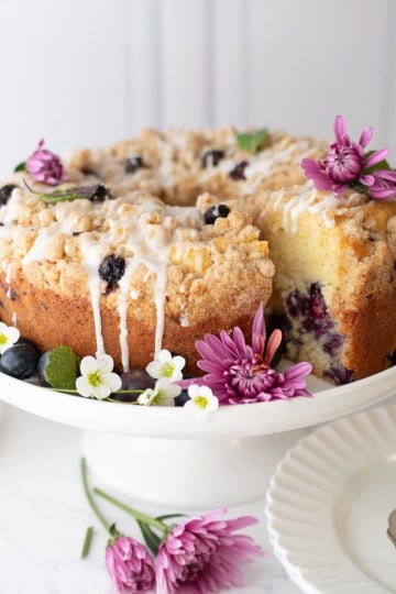 A blueberry yogurt teacake with icing is displayed on a white cake stand, garnished with fresh flowers and surrounded by plates and a bowl of blueberries.
