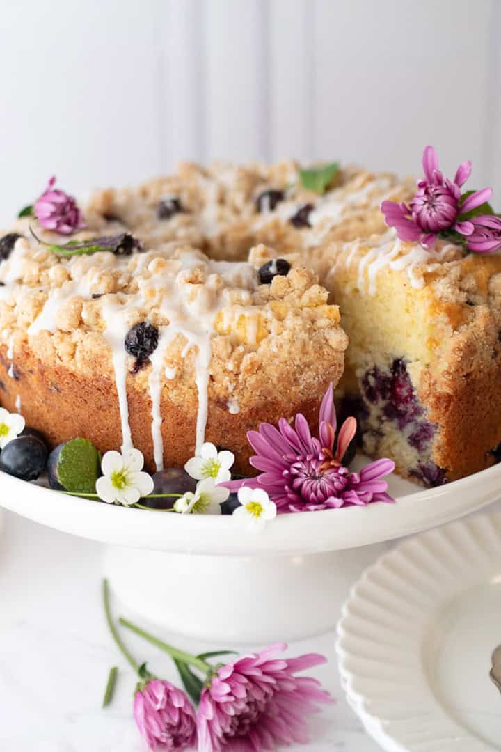 A blueberry yogurt teacake with icing is displayed on a white cake stand, garnished with fresh flowers and surrounded by plates and a bowl of blueberries.