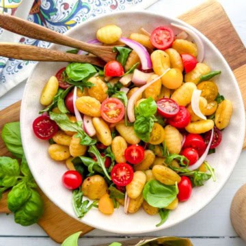 A bowl of gnocchi salad with cherry tomatoes, red onion, fresh basil, and arugula—perfect for easy dinners or spring dinners—served on a white plate with wooden utensils.