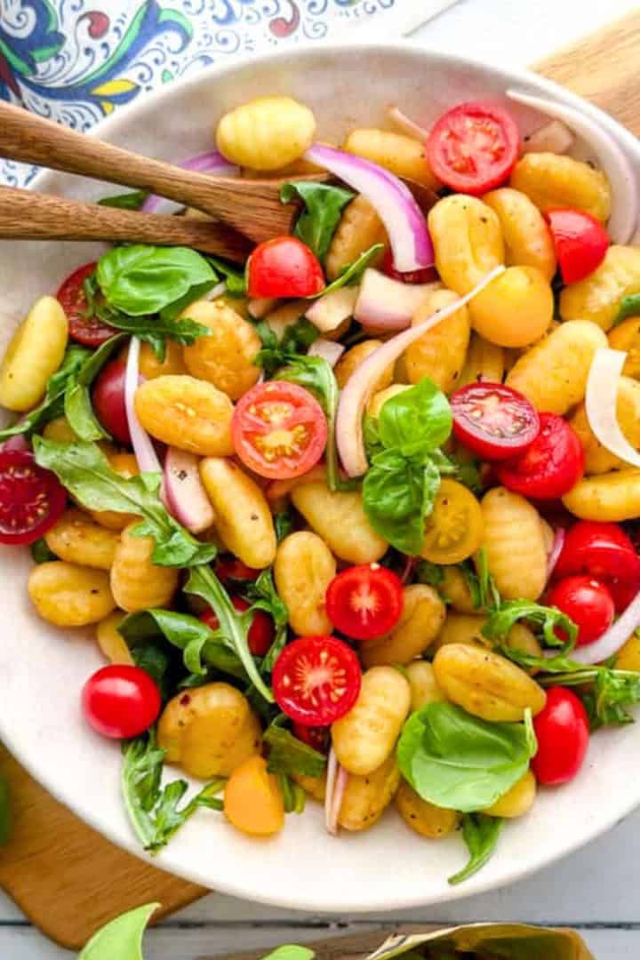 A bowl of gnocchi salad with cherry tomatoes, red onion, fresh basil, and arugula-perfect for easy dinners or spring dinners-served on a white plate with wooden utensils.