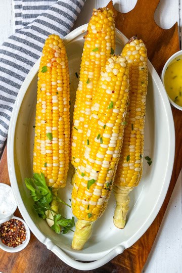 Four ears of corn on the cob with seasoning and herbs are served in a white dish, accompanied by small bowls of melted butter, salt, and red pepper flakes.