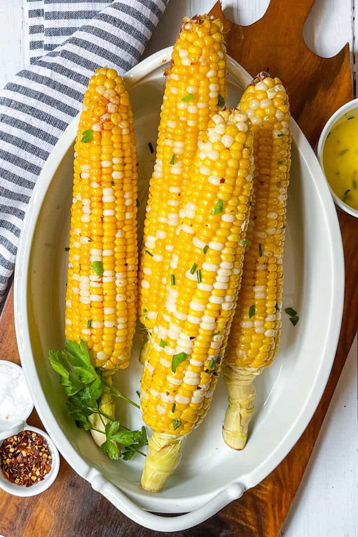 Four ears of corn on the cob with seasoning and herbs are served in a white dish, accompanied by small bowls of melted butter, salt, and red pepper flakes.