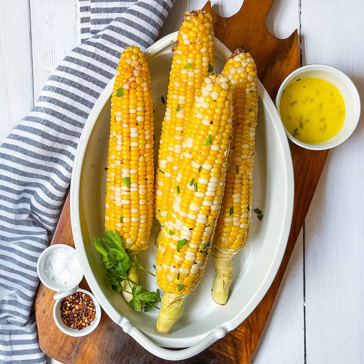 Four ears of corn on the cob with seasoning and herbs are served in a white dish, accompanied by small bowls of melted butter, salt, and red pepper flakes.