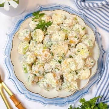 A plate of creamy Yukon Gold potato salad garnished with chopped green onions and parsley, placed on a decorative white and blue plate with gold utensils nearby.