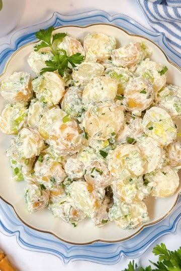 A plate of creamy Yukon Gold potato salad garnished with chopped green onions and parsley, placed on a decorative white and blue plate with gold utensils nearby.