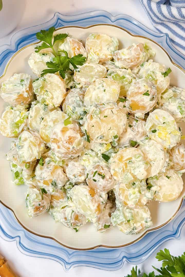 A plate of creamy Yukon Gold potato salad garnished with chopped green onions and parsley, placed on a decorative white and blue plate with gold utensils nearby.