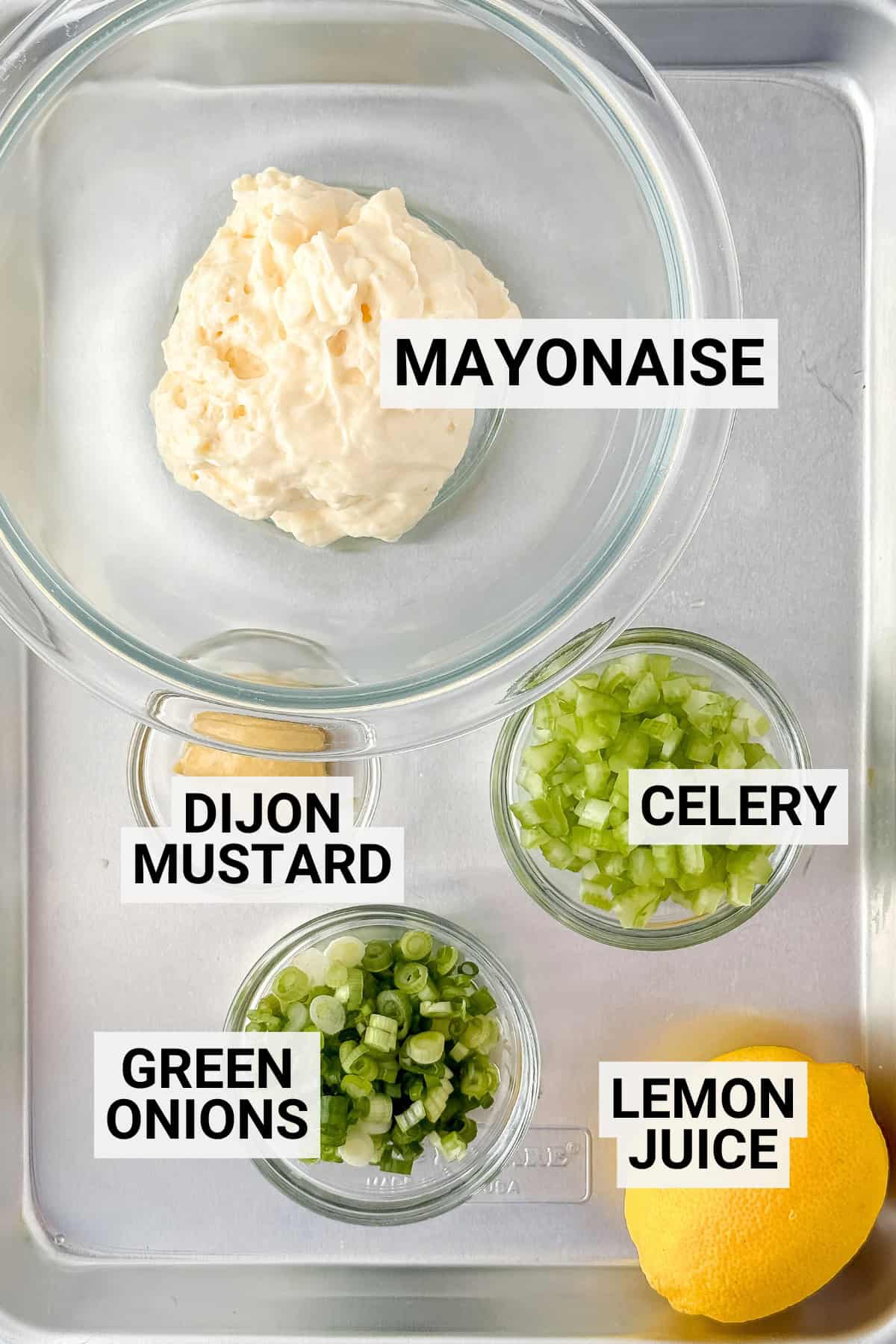 A tray with bowls of mayonnaise, chopped celery, chopped green onions, Dijon mustard, and a halved lemon-classic fixings for New England style shrimp rolls-each labeled with text.