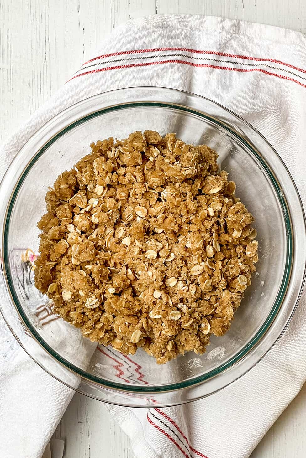 A glass bowl filled with an oat and crumb mixture for strawberry crisp sits on a white cloth with red stripes on a white wooden surface.