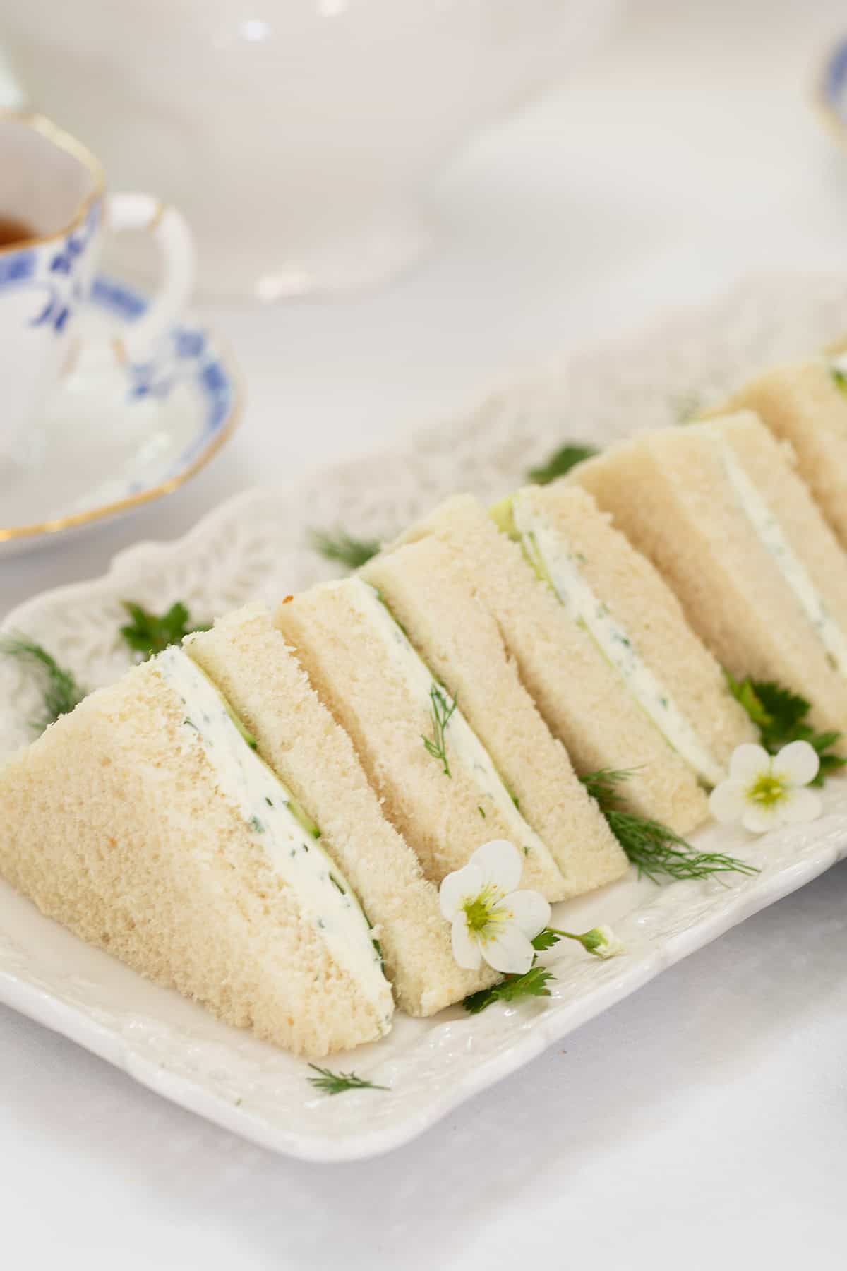 A rectangular white plate with several triangular Benedictine Tea Sandwiches, garnished with fresh dill and small white edible flowers.