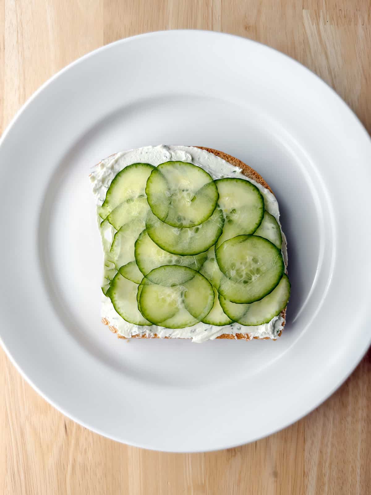 A slice of bread with cream cheese and overlapping cucumber slices on a white plate, set on a wooden surface-perfect for classic Kentucky Benedictine tea sandwiches.
