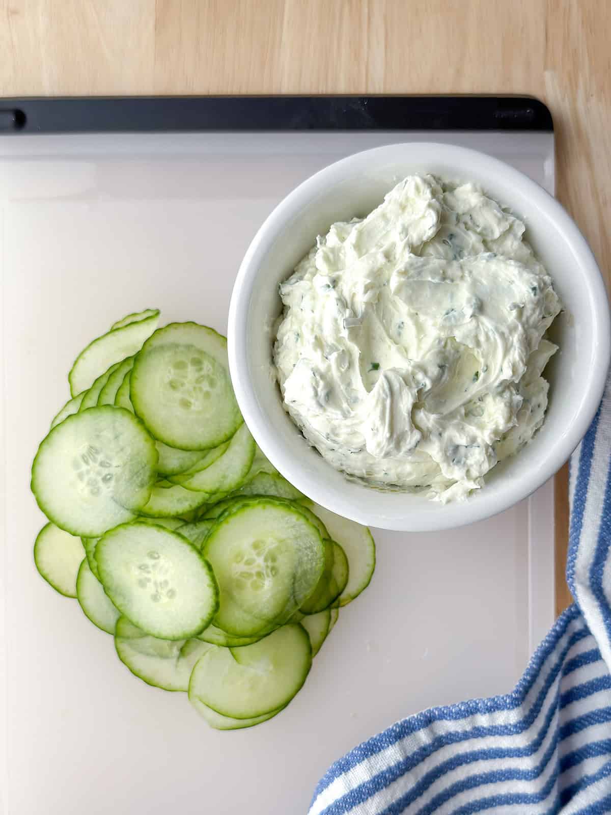 A bowl of herbed cream cheese sits next to neatly sliced cucumber rounds on a white cutting board-perfect for assembling classic Kentucky Benedictine tea sandwiches, with a striped towel nearby.