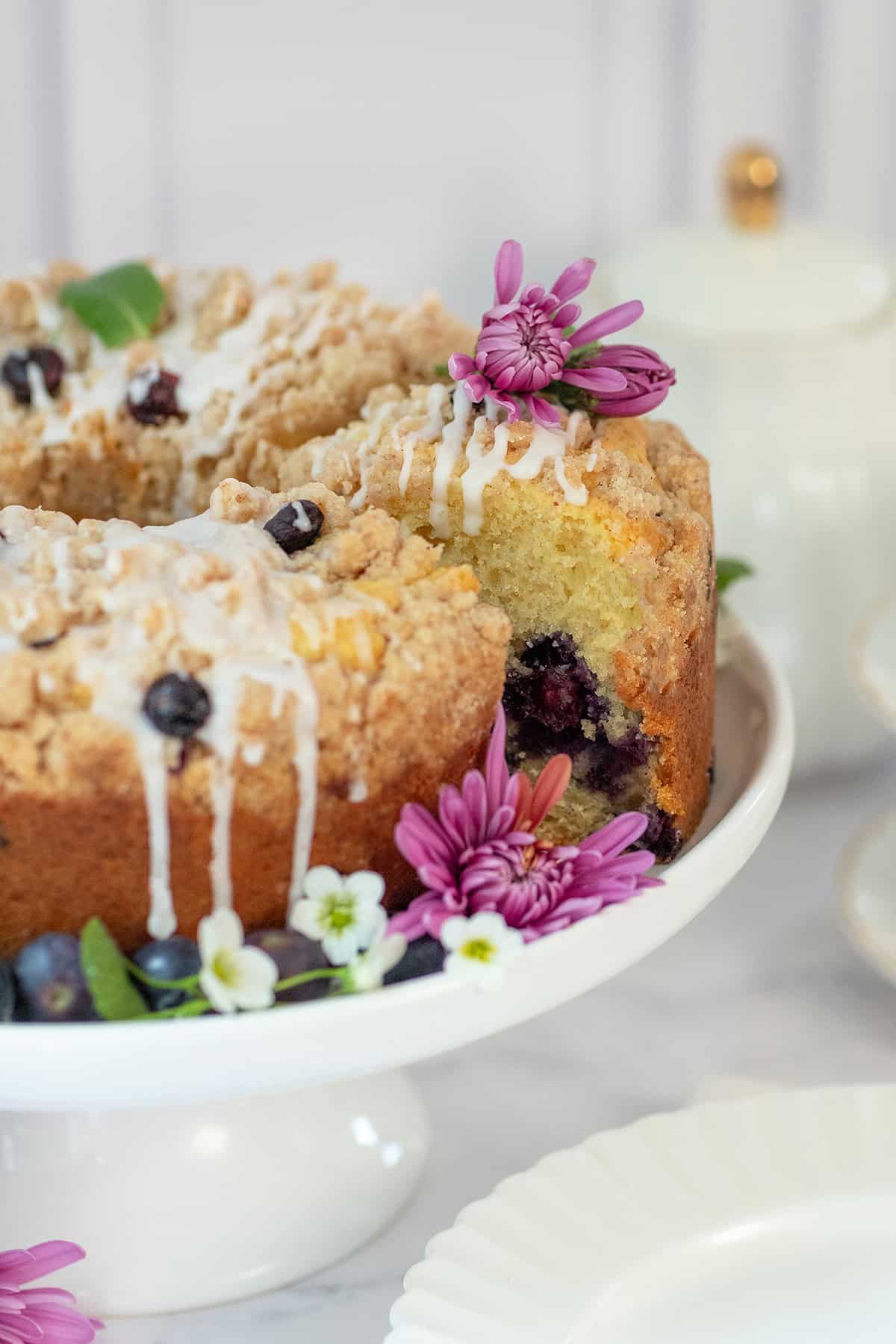 A blueberry yogurt teacake with icing and crumb topping sits on a white cake stand, adorned with purple flowers and green leaves. A teapot and plates are in the background.
