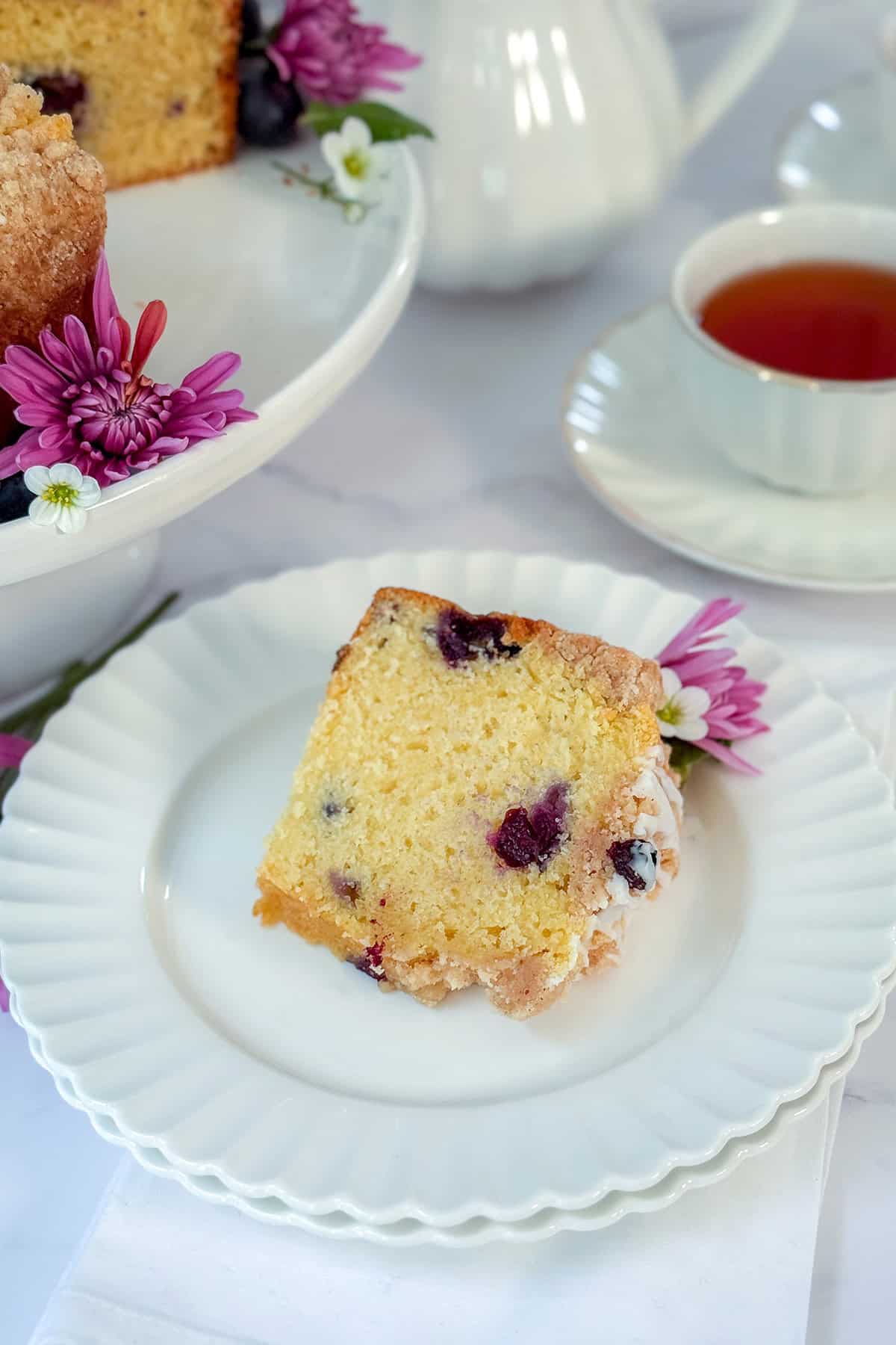 A slice of blueberry yogurt teacake on a white plate, garnished with purple flowers, with a cup of tea and a cake stand in the background.