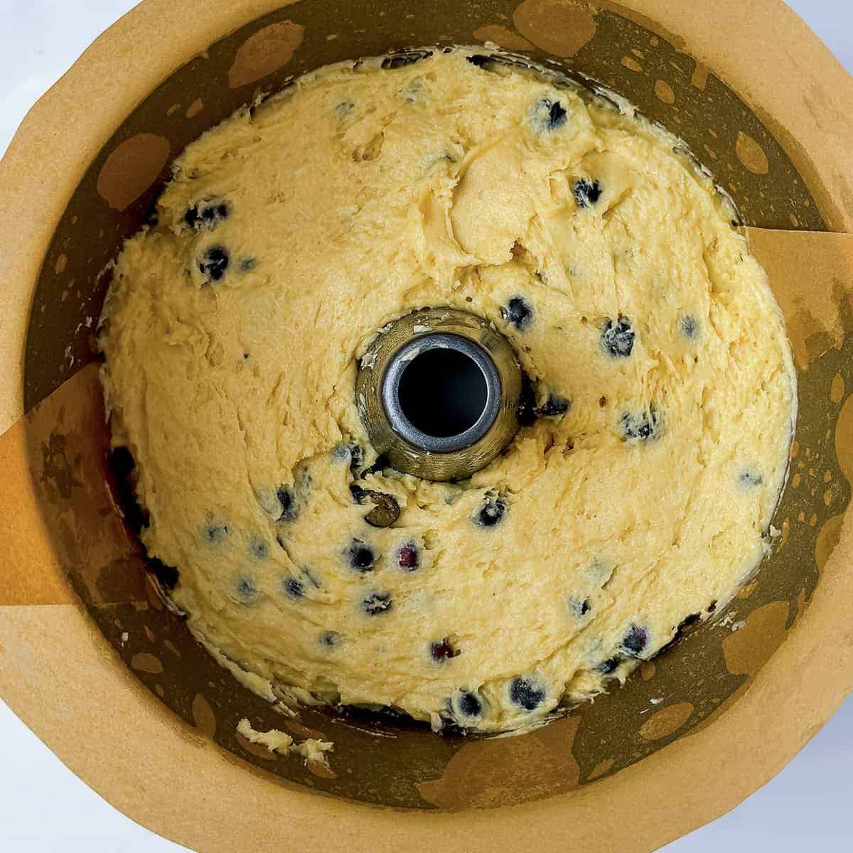 A bundt cake pan lined with parchment paper, filled with unbaked blueberry and yogurt teacake batter, sits on a marble surface.