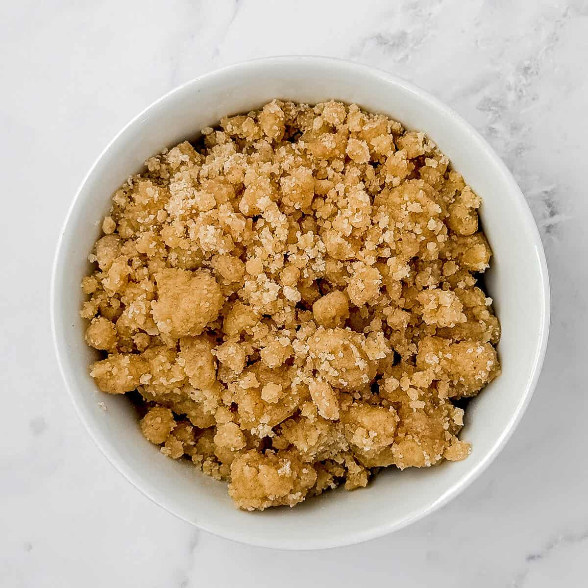 A white bowl filled with crumbly brown sugar streusel, perfect for topping a blueberry teacake, sits on a white marble surface.