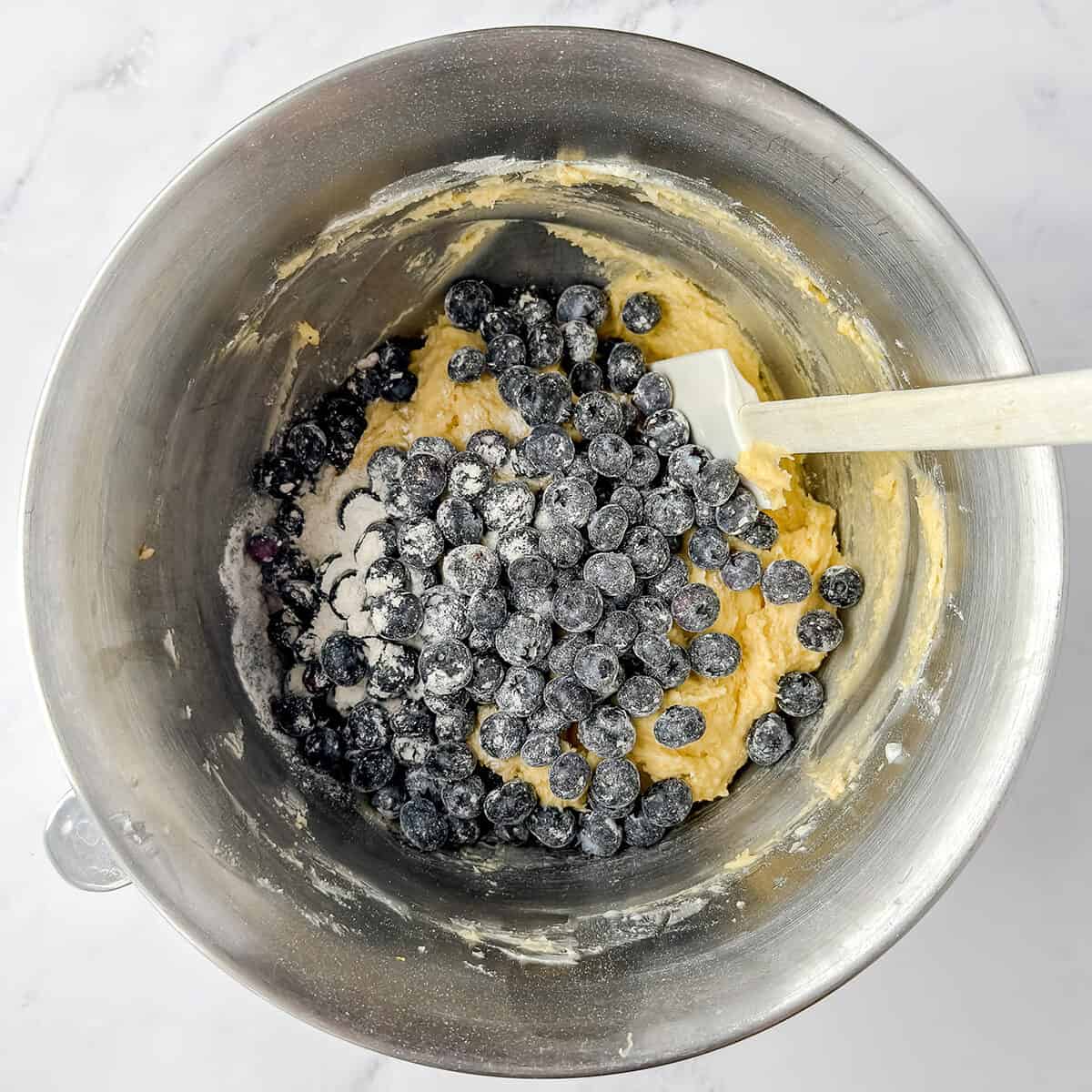 A metal mixing bowl containing yellow teacake batter, fresh blueberries, and a white spatula resting inside, on a white marble surface.