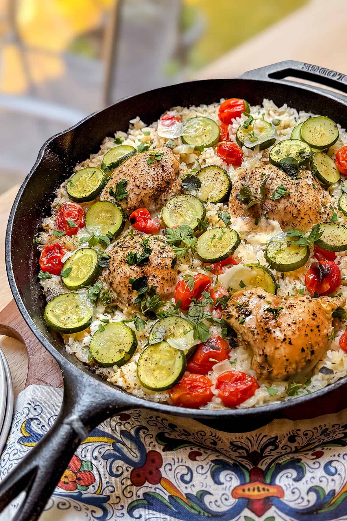 A one pan cast iron skillet with Tuscan chicken thighs, sliced zucchini, cherry tomatoes, and rice, garnished with fresh herbs, rests on a patterned cloth.