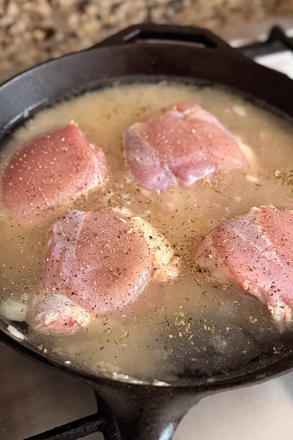 Four raw chicken thighs are simmering in a cast iron skillet filled with broth, seasoned with black pepper and herbs for a flavorful one pan dinner.