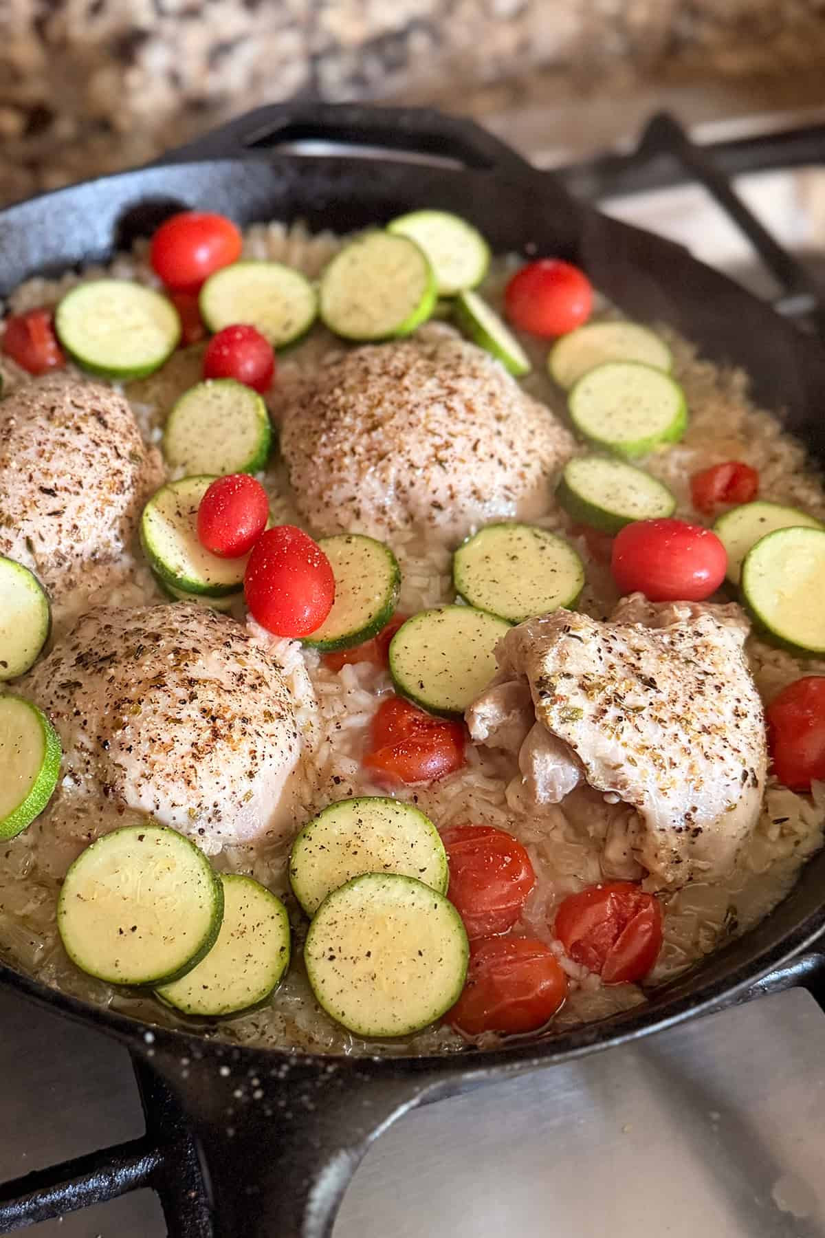 Chicken thighs, zucchini slices, cherry tomatoes, and rice cook together in a cast iron skillet on the stovetop.