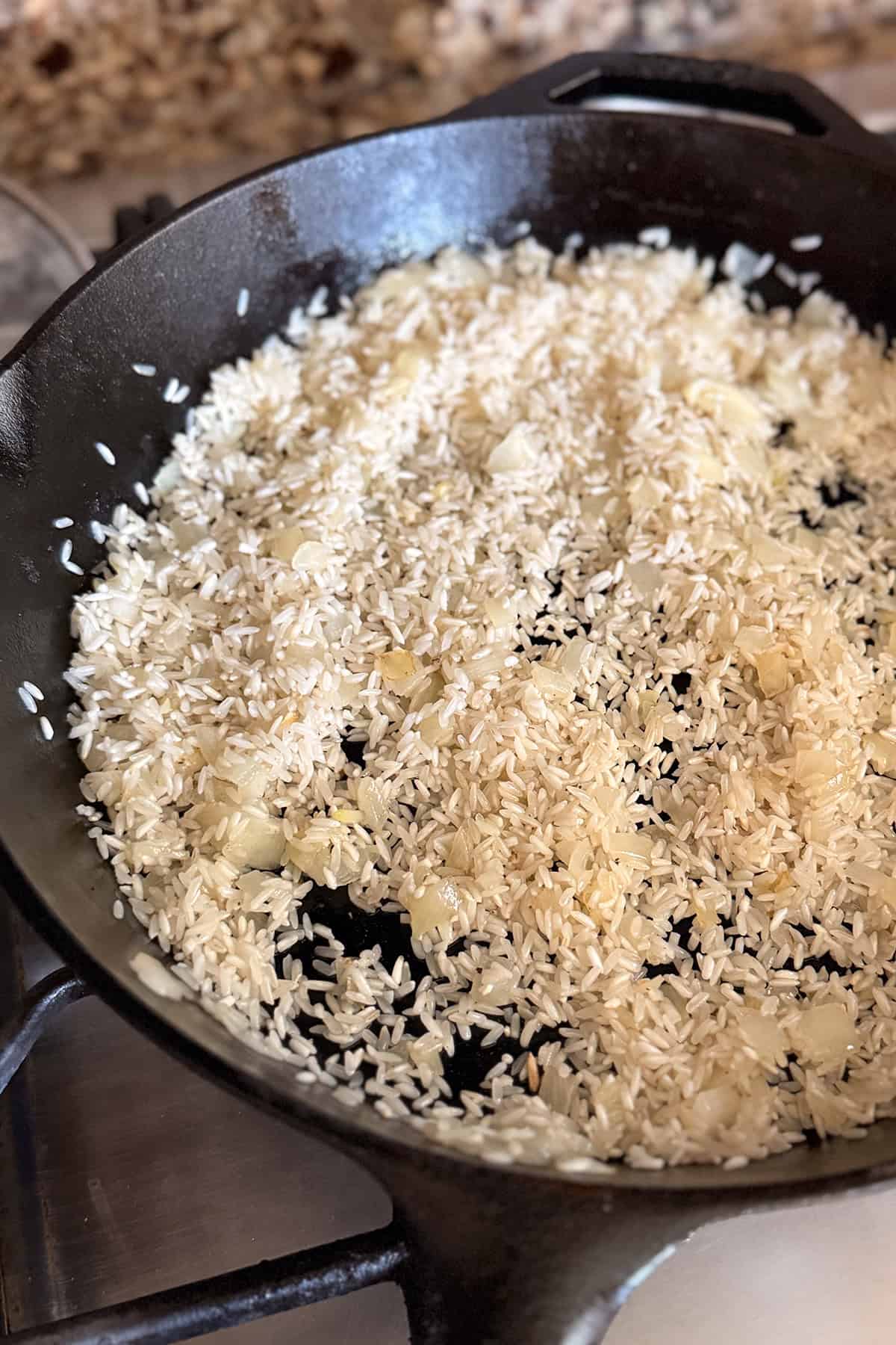 Uncooked rice and chopped onions are being saut&eacute;ed in a black cast iron skillet on a stove, creating the perfect base for a flavorful one pan skillet chicken and rice dish.