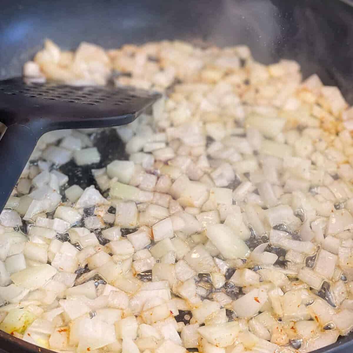 Diced onions being saut&eacute;ed in a nonstick frying pan with a black spatula on a stovetop, perfect for starting a classic German Potato Salad.