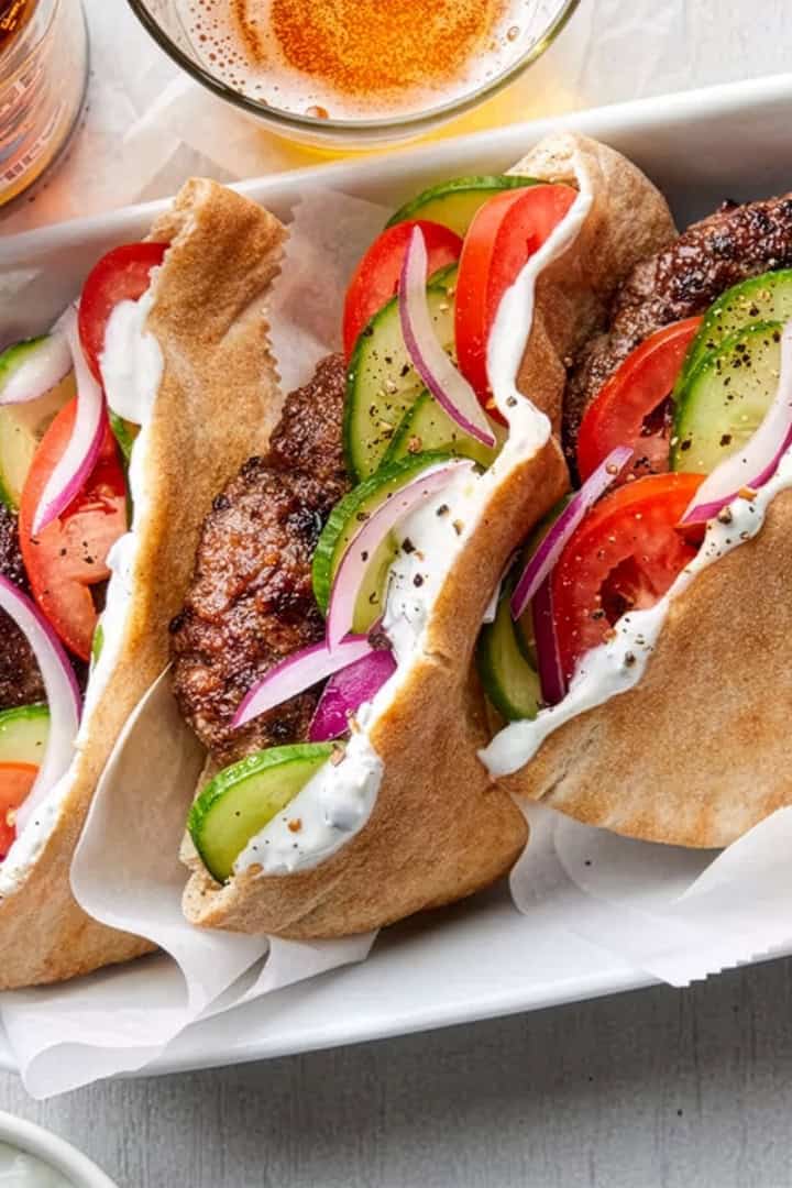Three pita sandwiches filled with seasoned meat, sliced cucumbers, tomatoes, red onions, and a dollop of white sauce-an easy May one pot dinner-served in a tray with drinks visible in the background.
